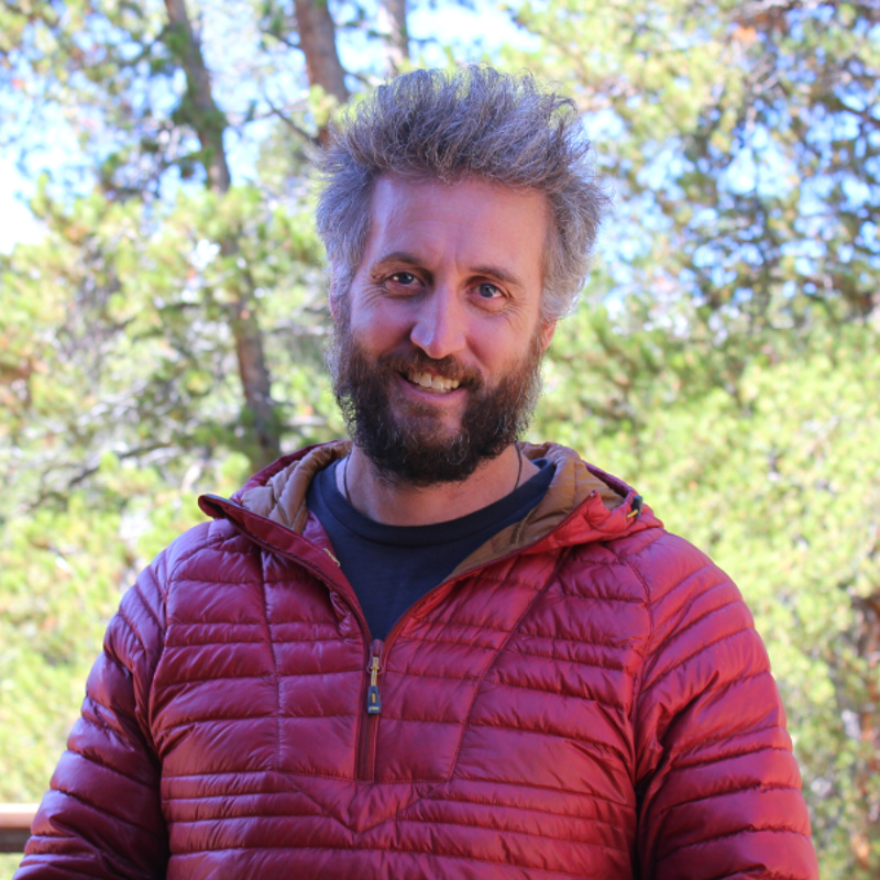 The image shows a man with a beard and graying hair, smiling at the camera. He is wearing a red puffy jacket with a hood. The background features trees and foliage, suggesting an outdoor setting. The man appears to be in a natural environment, possibly a forest or park.